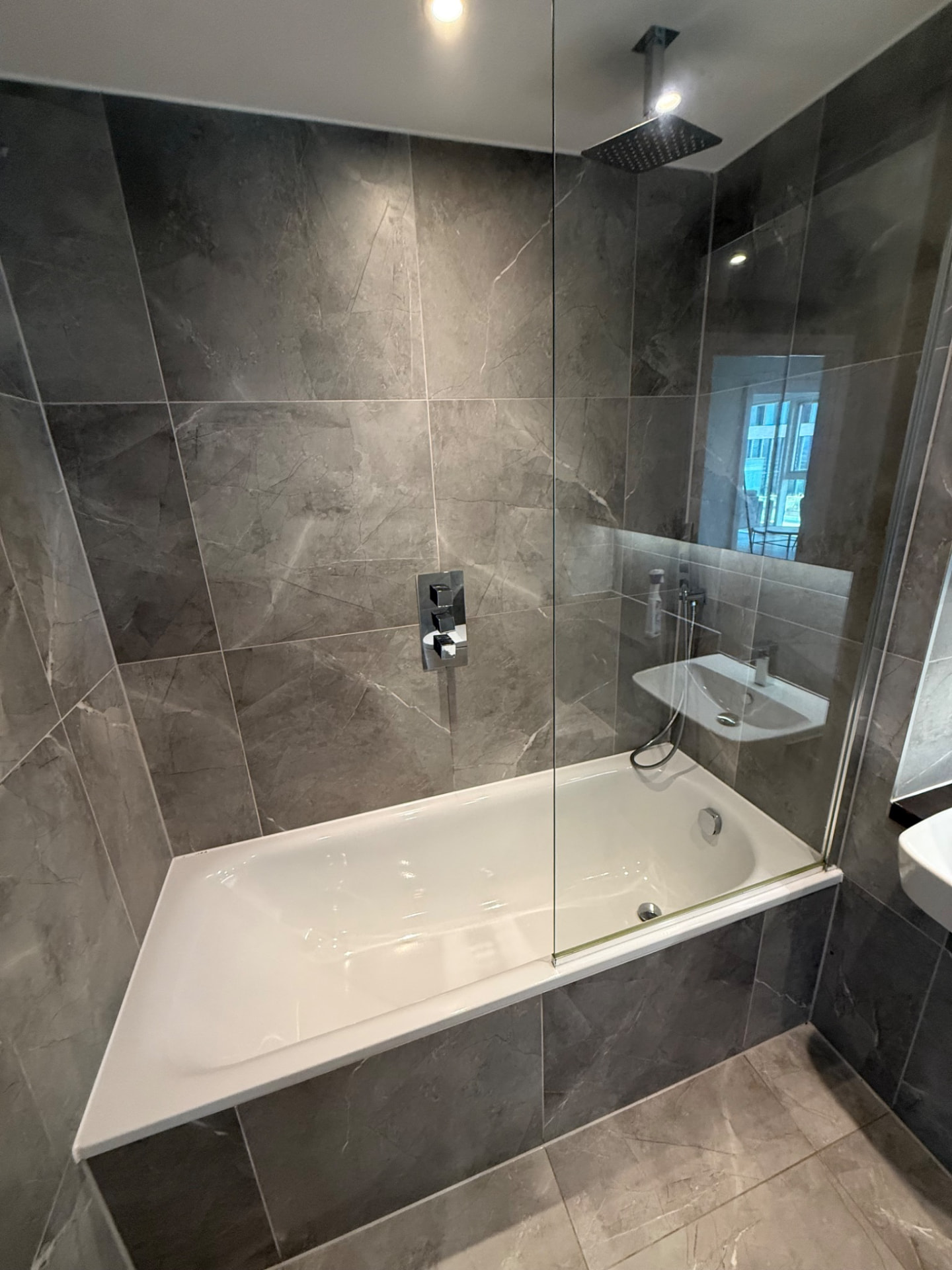 Bath with a glass shower screen and grey tiled walls